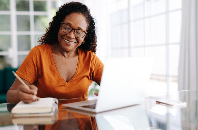 Happy senior woman sitting at a table in her home office, drafting her last will and testament in a journal and taking care to allocate her assets. Retired woman leaving a plan in place for the future.