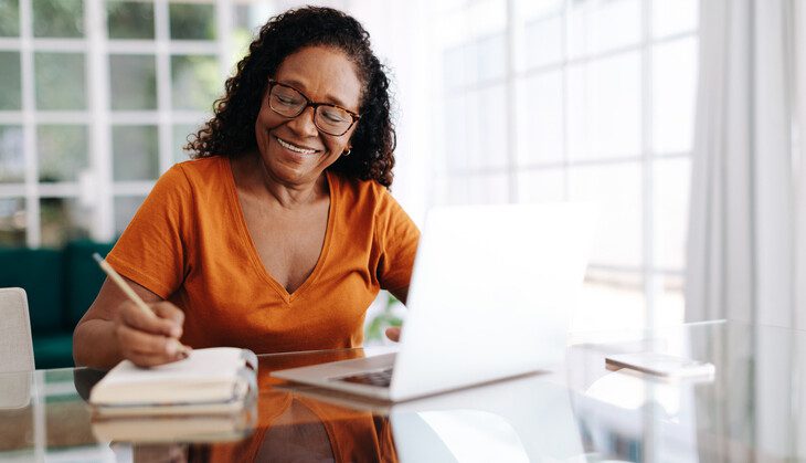 Happy senior woman sitting at a table in her home office, drafting her last will and testament in a journal and taking care to allocate her assets. Retired woman leaving a plan in place for the future.